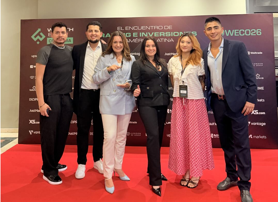 Six people posing on a red carpet in front of a branded event backdrop with logos behind them.