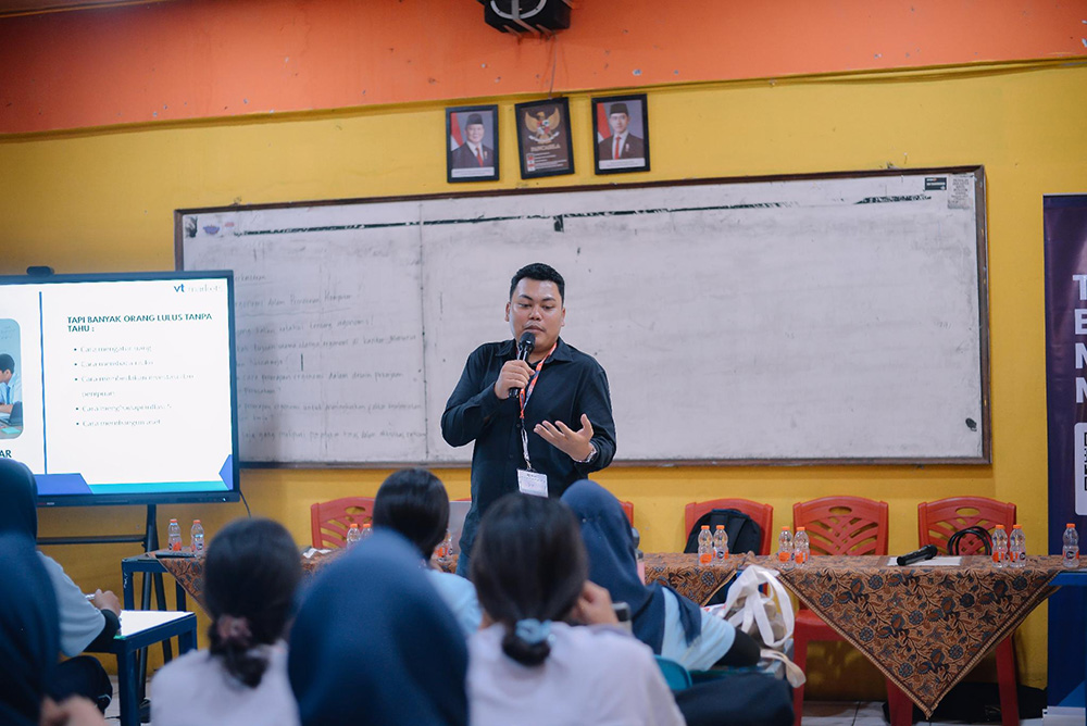 A man speaks into a handheld microphone at a classroom presentation, addressing seated students with a slide on the screen to the left.