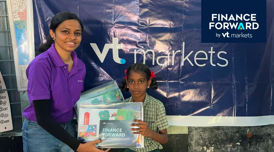 Woman in a purple polo hands a stack of vt markets finance materials to a young girl beside a large vt markets banner and Finance Forward logo.